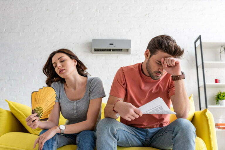 man with newspaper and woman with hand fan sitting on sofa and suffering from heat at home
