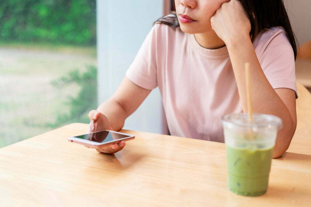 Bored young asian woman looking disappointed at her smart phone while waiting someone in restaurant