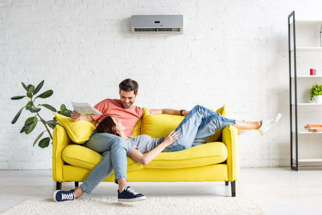 happy man with smiling girlfriend relaxing on yellow sofa under air conditioner at home